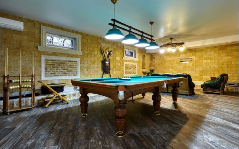 Pool table in a basement with small windows with a brick façade wall covering and diagonal grey wood-look tile