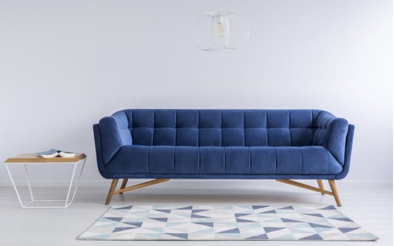 Living room with a simple blue velvet couch next to a modern white end table with a light wooden top and a triangle patterned rug in various shades of blue and white