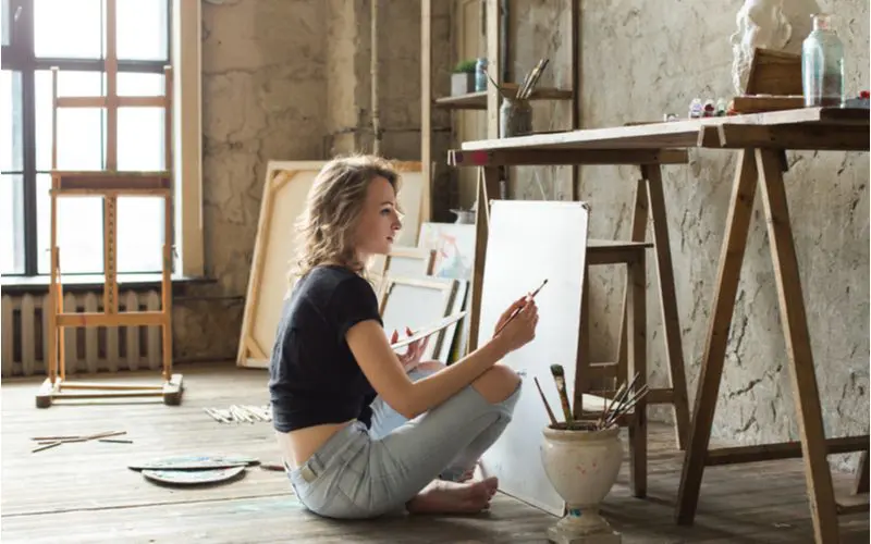 For a piece on what type of paint to use on picture frame, a woman sitting cross legged on the floor painting a frame