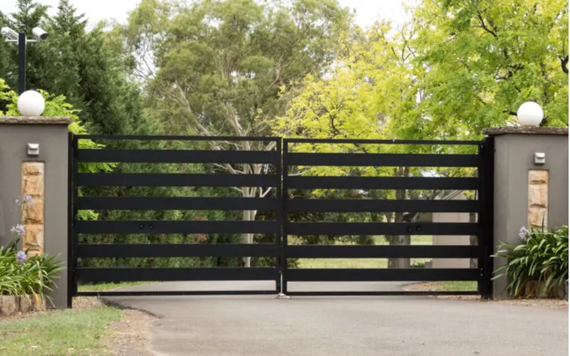 Modern metal driveway gate idea featuring a horizontally slatted black metal driveway gate between two stucco supports with round lights on top of each