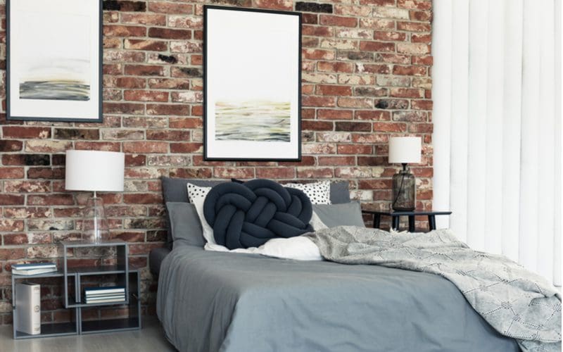 Photo of a men's bedroom idea showing how easily a natural red brick wall can be spruced up with a few black framed posters, a some modern geometric nightstands, and a grey comforter