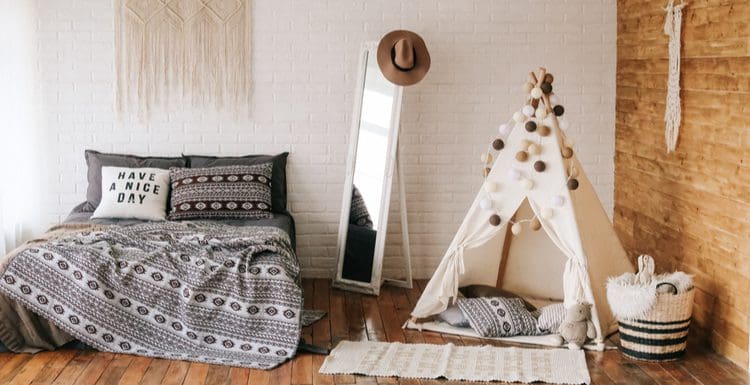 Featured image for a piece titled Boho Bedrooms featuring a small white teepee, lots of macramé tapestries and blankets, and a bohemian-style hat hanging from a large freestanding mirror