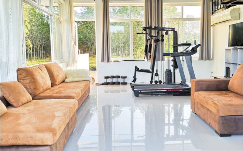 Small home gym with polished white concrete floors and beige couches next to a bowlex