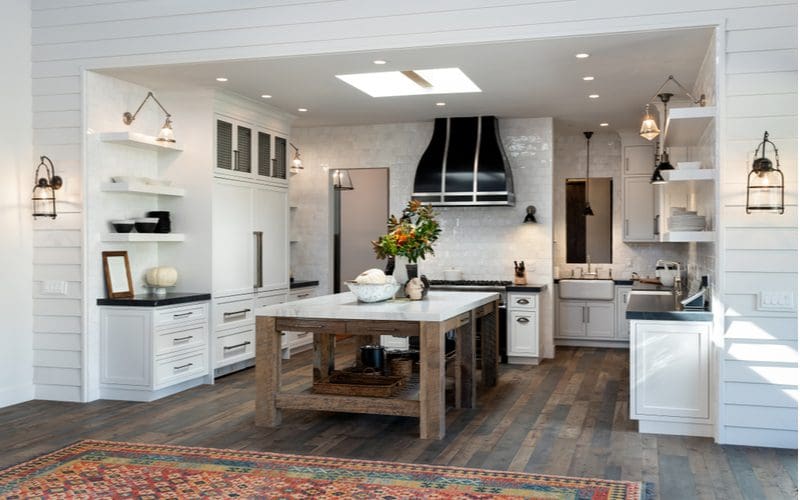 Kitchen that has a farmhouse island made of grey stained timber, rustic white painted cabinets, white shiplap walls, and dark countertops and fixtures