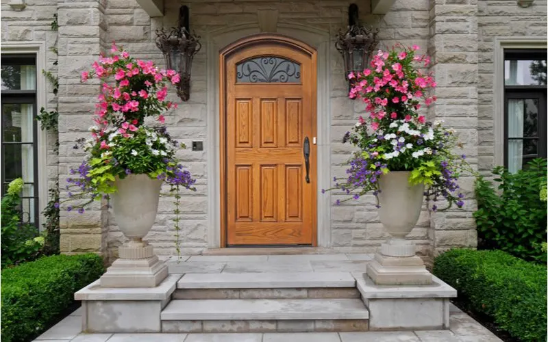 Trophy-Shaped entryway planter ideas made of stone and sitting on either side of the stone square paver walkway leading to the front door made of natural wood