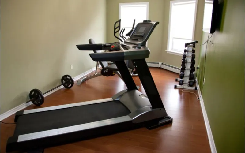 Small home gym with a dumbell rack and treadmill and elliptical below a tv with two big windows with ledges in a taupe room