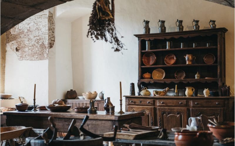 European gothic style kitchen with rustic cabinets that are stained dark brown with white stucco walls with natural brown pots sitting on top