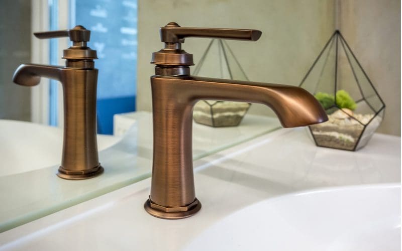 Copper single hole faucet reflected in the mirror above a farmhouse-style porcelain sink bowl to give a modern rustic look to a bathroom