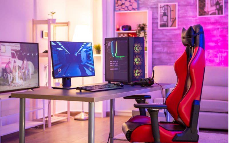 Image for a piece on gaming setup ideas featuring a red chair in front of a simple wooden slab desk with metal legs on which two monitors sit with purple lighted walls in the background