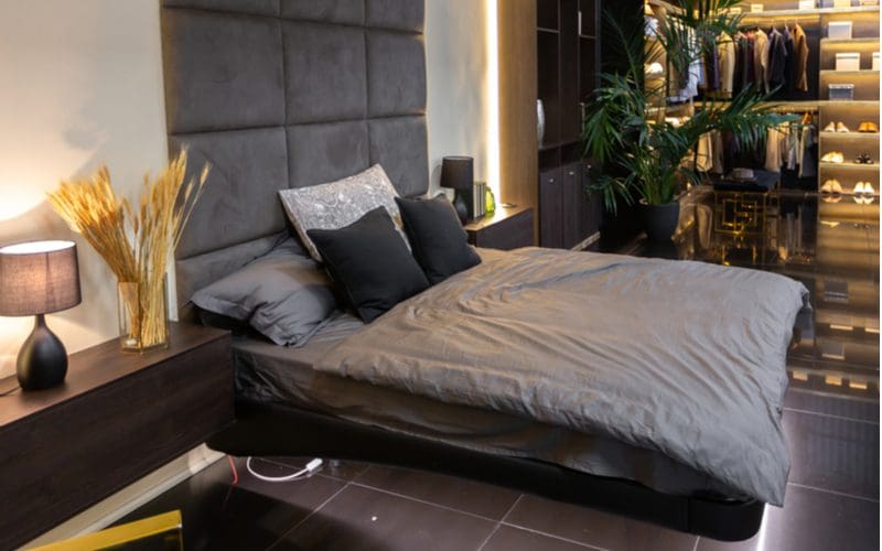 Bachelor pad idea featuring a floating black bed above dark grey ceramic tile and a grey upholstered headboard attached to the wall with a large open closet in which you can see shoes, shirts, and more