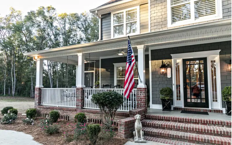 Image showing which side of porch to hang American flag on, as depicted on a large Colonial-style home that's painted grey with ample red brick