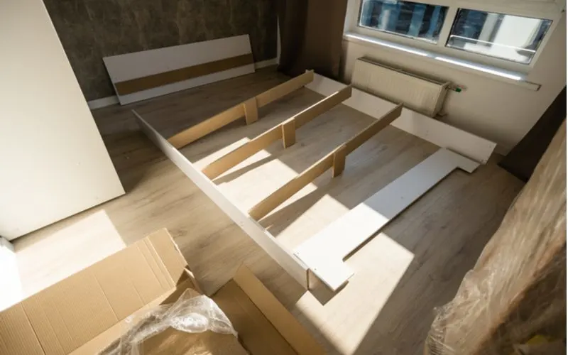Various parts of a bed laid out on a grey wood-look laminate floor in a small studio apartment