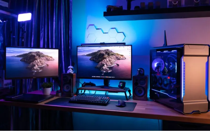 For a piece on gaming setup ideas, an ironman-type PC sits on a wooden desk with two monitors and a speaker behind a blue ambient wall with white hex tiles on it