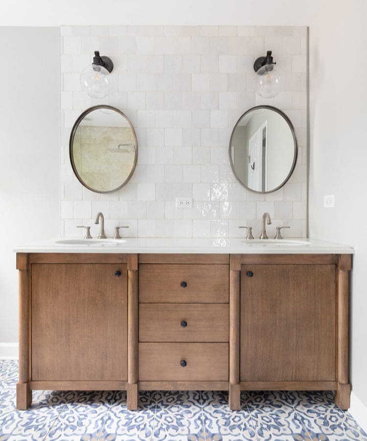 As an idea for modern rustic bathroom ideas, a bunch of Spanish-style floor tile holds up a rustic wooden vanity with twin round mirrors below streelight-style lights