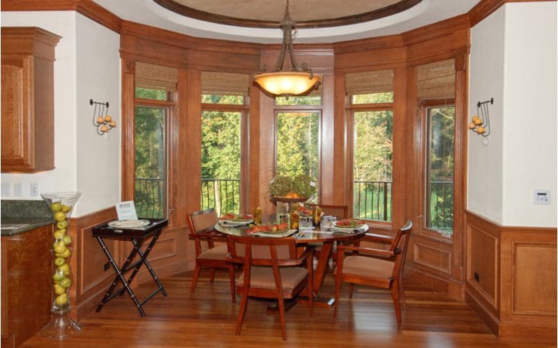 For a piece on breakfast nook ideas, a round-walled dining area with padded chairs and a serving tray overlooks a wooded backyard