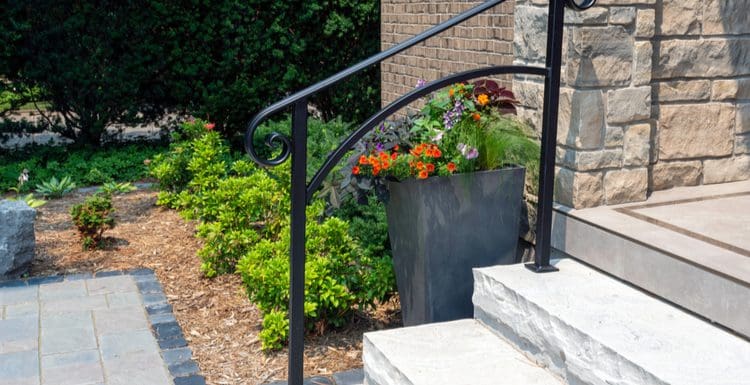 Featured image for a design roundup titled Entryway Planter Ideas featuring a square stone flower planter in a cedar mulch landscaping bed next to a concrete staircase with a black railing on it
