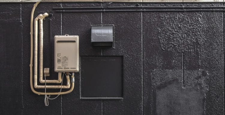 Featured image for a piece titled can you put a water heater outside depicting such an appliance in the tankless variety outside and attached to a black wall