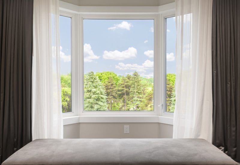 Bay window curtain idea featuring a scenic landscape framed by white bay windows with brown blackout and white sheer curtains, both drawn