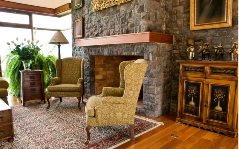 Vintage style cottage interior with plush and ornate chairs on a fancy rug next to a stone mantle and fireplace wall