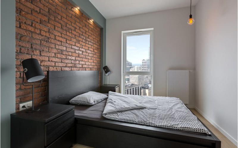 Bedroom with industrial styling has a brick wall, hanging Edison bulb light fixture, a desk lamp with a black wire frame, and a small window that overlooks a city