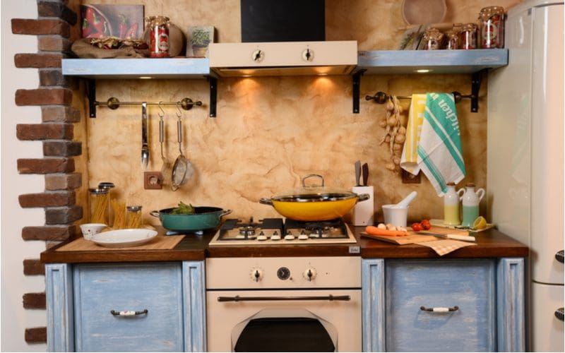 Rustic cabinets painted blue and purposely weathered hold up a butcher block countertop stained dark in color with a brick and stucco wall next to the stove