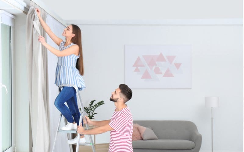 Image included for a piece on how to hang curtains without nails featuring a woman standing on a step ladder with a guy holding it while she hangs the curtains