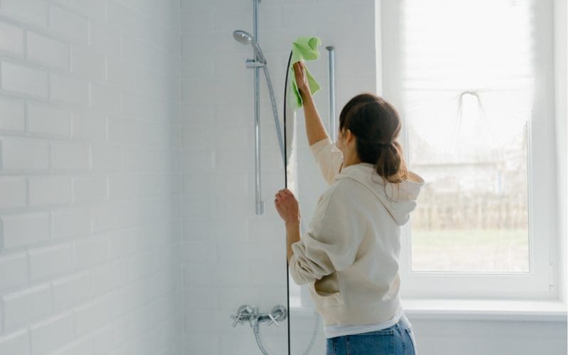 Woman cleaning a fiberglass shower using a microfiber rag and cleaning products while wearing jeans and a hoodie