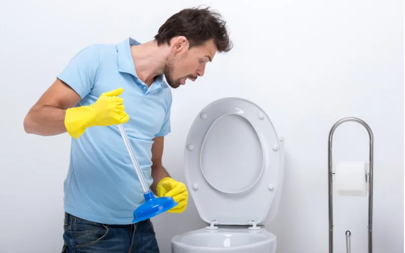 Image of a man in a blue shirt showing us how to unclog a toilet with poop in it with a small blue plunger and gloved hands