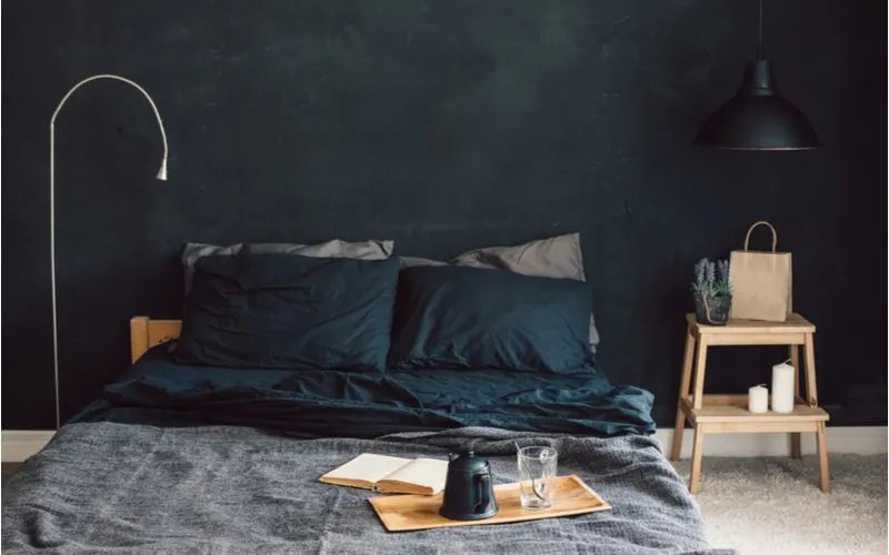 Room idea with black walls and dark charcoal and grey bedding with a wooden table lamp next to the bed below a hanging black lamp