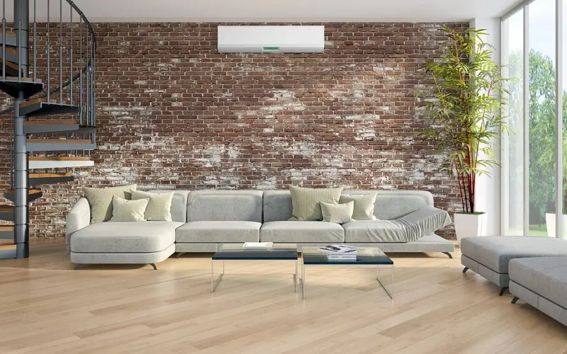 Large light gray sofa with brown brick wall behind and large windows 