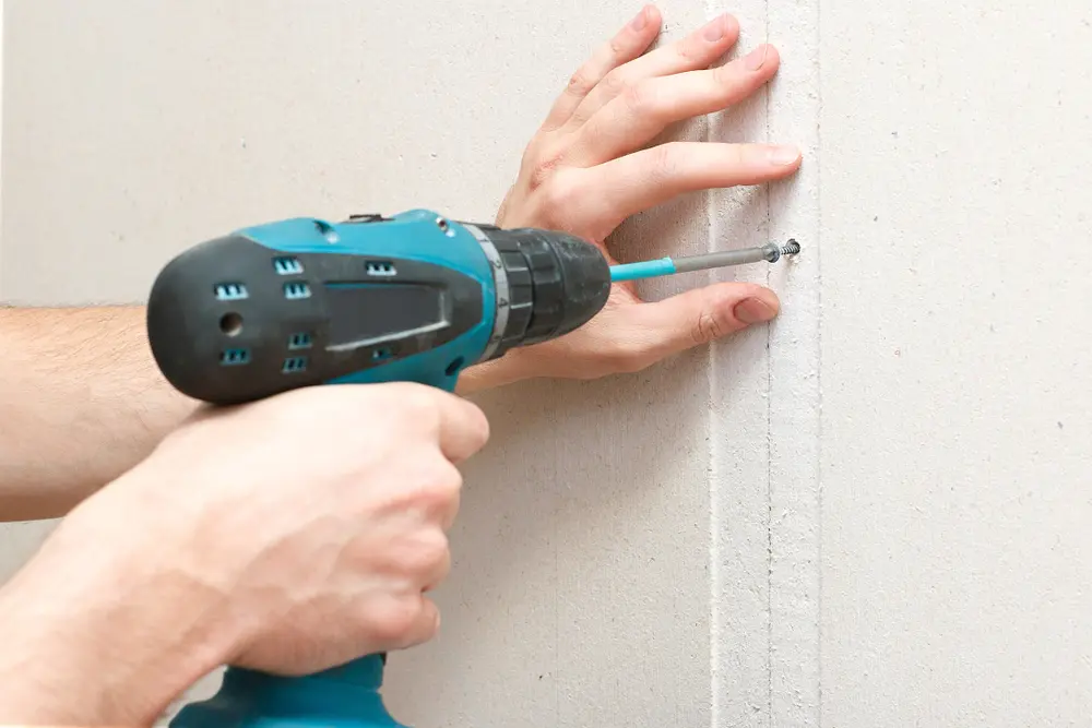 Guy with a Makita drill in teal color using one of the standard sized drywall screws to attach a piece of drywall to the wall