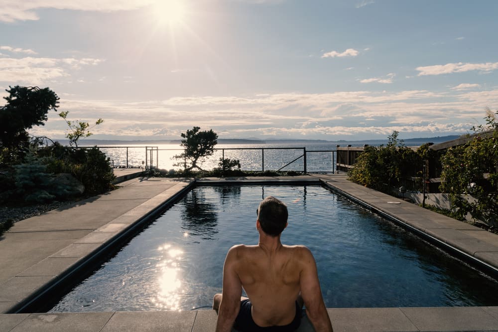 Man in a backyard sitting next to an average-sized lap pool as the sun sets over the ocean