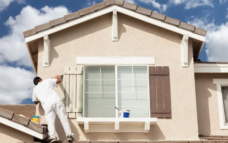 Man painting shutters in cream color on a tann house exterior