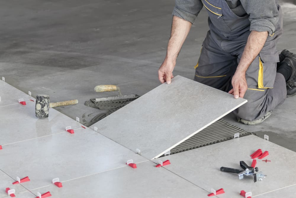 Ceramic tile, one of the best types of tile for flooring, being laid by a guy in work pants and wearing kneepads